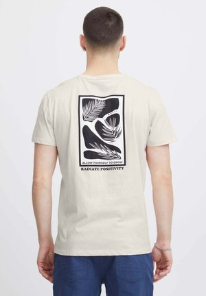 T-shirt in cotone beige con una grafica in bianco e nero sul retro, con motivi di foglie di palma e le frasi: "PERMETTI A TE STESSO DI CRESCERE" e "RADIATE POSITIVITÀ."