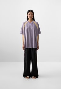 Una donna si trova di fronte, indossando una blusa viola chiaro ampia con dettagli gialli e pantaloni neri a gamba larga. Ha i capelli neri lunghi e lisci.