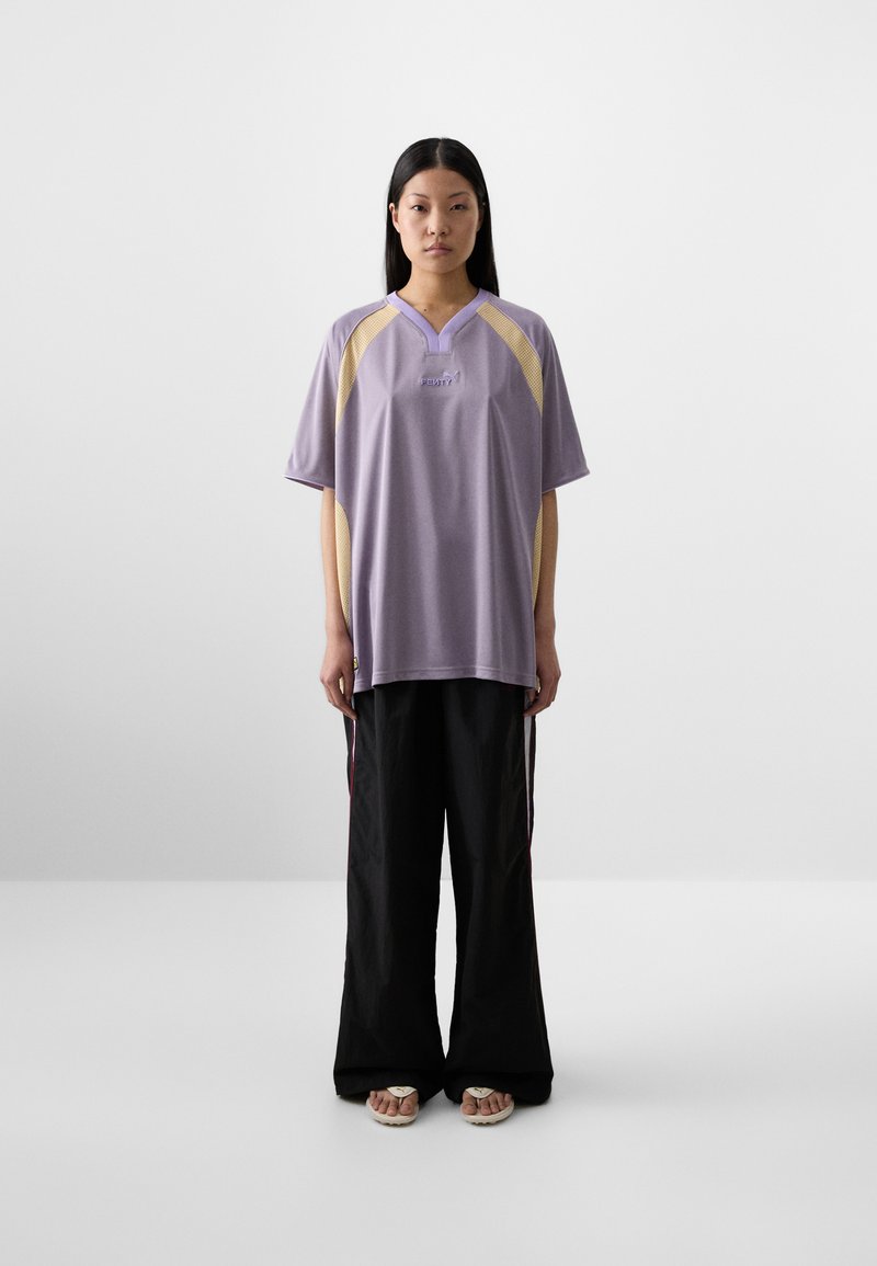 Una donna si trova di fronte, indossando una blusa viola chiaro ampia con dettagli gialli e pantaloni neri a gamba larga. Ha i capelli neri lunghi e lisci.