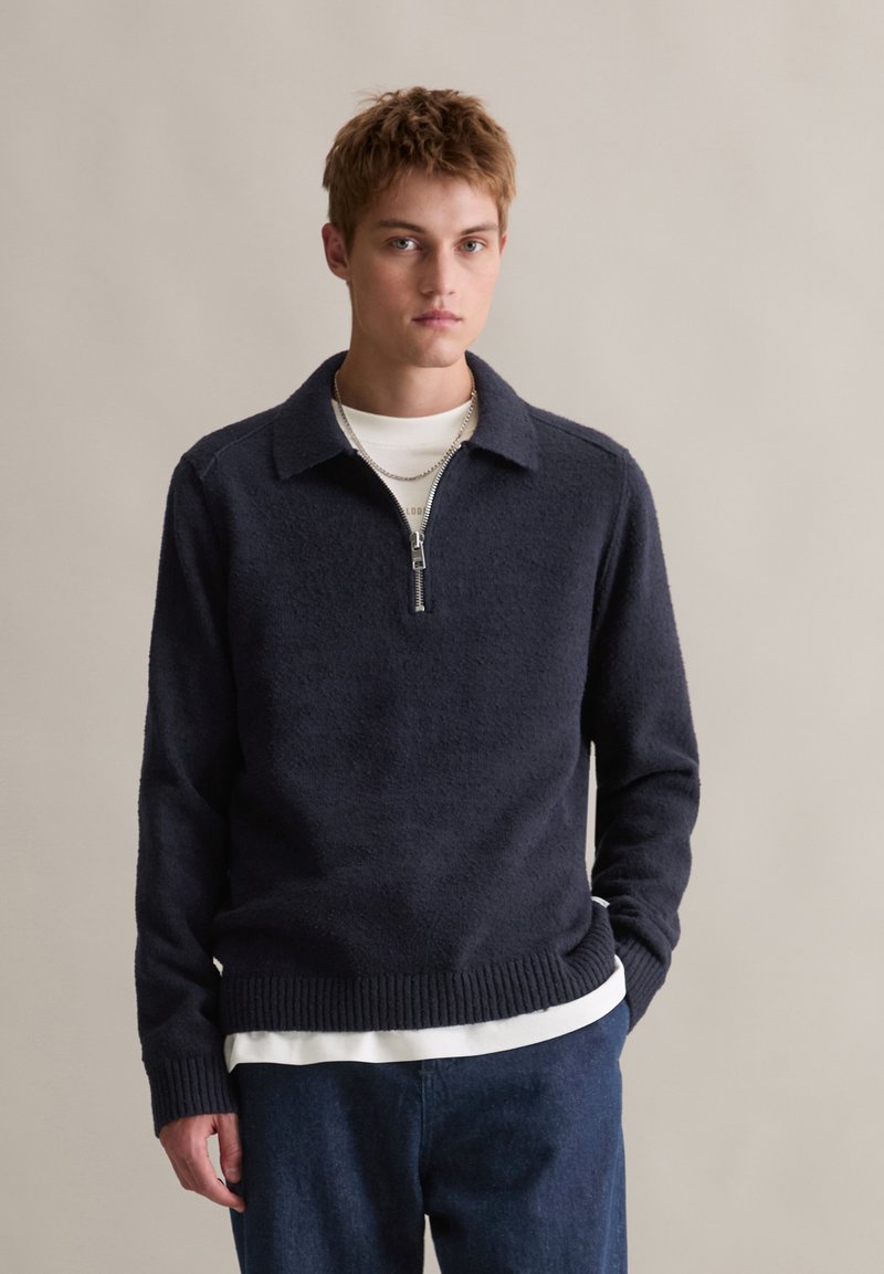 Granatowy sweter z dzianiny z ćwierć zapięciem i kołnierzykiem. Posiada teksturowany materiał i ściągacz u dołu. Noszony na białej koszuli z dżinsami.