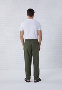 Vit t-shirt med rund hals och olivgröna byxor med elastisk midja och två bakfickor, kombinerat med svarta sandaler. Modellen står mot en tegelvägg.