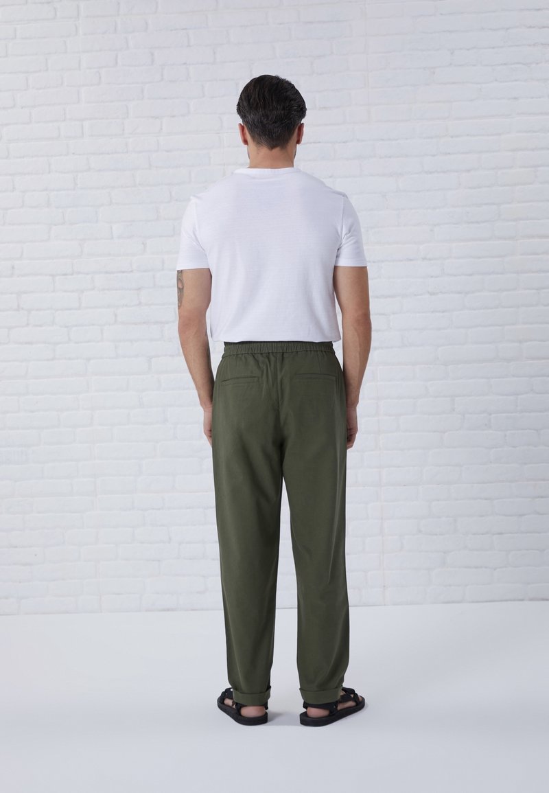 Vit t-shirt med rund hals och olivgröna byxor med elastisk midja och två bakfickor, kombinerat med svarta sandaler. Modellen står mot en tegelvägg.