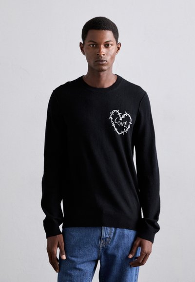 Homme portant un sweater noir avec un design de fil barbelé en forme de cœur blanc et le mot "AMOUR", associé à un jean bleu, se tenant devant un fond uni.