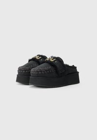 Čierne platformové slip-on topánky s kožušinovým akcentom, ozdobným prešívaním a zlatým logom na pásku. Ploché gumové podrážky.