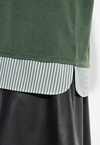 Felpa verde con orlo arrotondato, indossata sopra una camicia a righe bianche e verdi con colletto; il tessuto in pelle nera è visibile sotto.