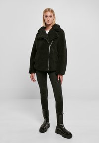 Zwarte nepbontjas met een brede kraag, ritsluiting en zijzakken, gecombineerd met zwarte slim fit broek en stevige zwarte laarzen.