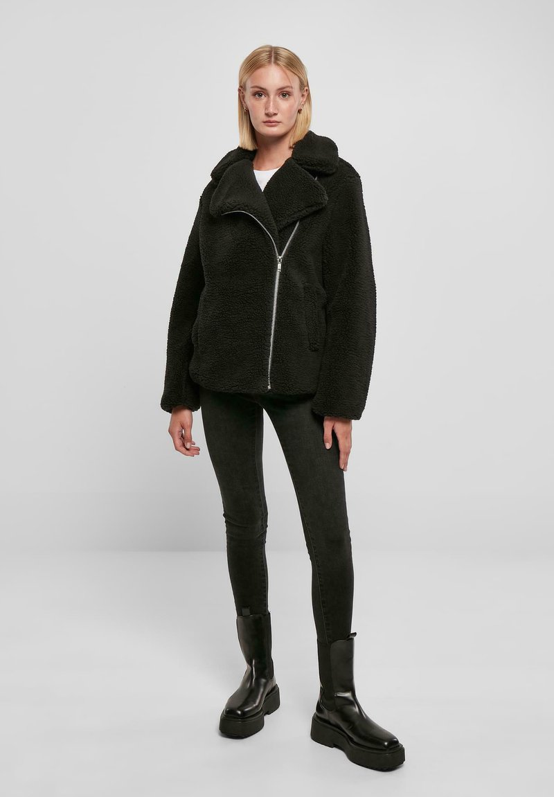 Zwarte nepbontjas met een brede kraag, ritsluiting en zijzakken, gecombineerd met zwarte slim fit broek en stevige zwarte laarzen.