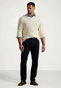 Vīrietis, kuram mugurā ir krēmkrāsas adītas džemperis virs zila krekla, tumšas džinses un brūni loferi, stāv uz vienkārša gaiša fona.