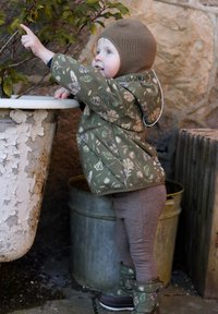 Pikowana zielona kurtka w kwiatowe wzory, szare legginsy i ocieplane buty. Dziecko bawi się przy roślinie w teksturowanej doniczce.