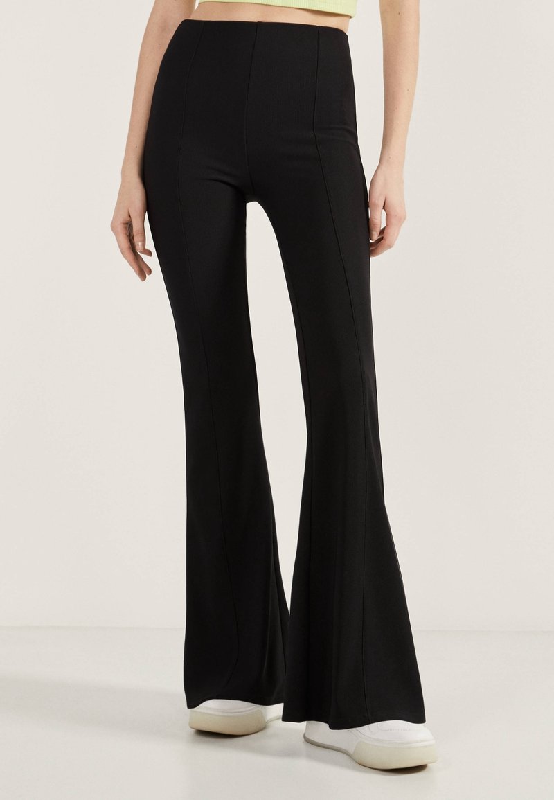 Svarta utsvängda byxor i ett stretchigt material, med hög midja och vertikala sömmar, matchat med vita sneakers. Slät textur.