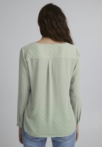 Blouse vert clair avec des pois blancs, manches longues, détail de empiècement au dos et ourlet arrondi. Fabriquée en tissu lisse et léger.