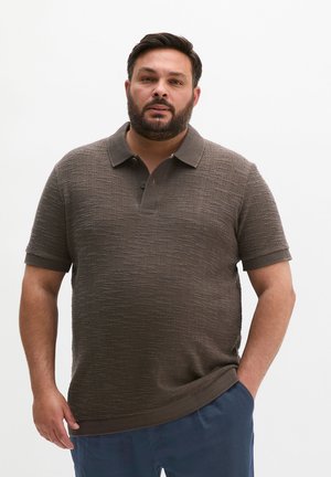 Mężczyzna z brodą, ubrany w brązową, teksturowaną koszulkę polo z krótkim rękawem i ciemnoniebieskie spodnie, stoi z jedną ręką w kieszeni na białym tle.