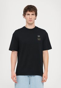Black cotton t-shirt with a round neckline, short sleeves, and a small logo patch featuring gold accents on the chest.