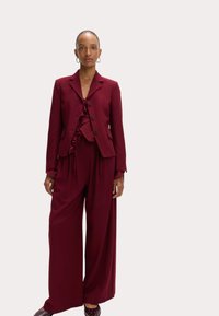 Traje burdeos a medida con una blusa con volantes, que incluye un blazer abotonado y pantalones de pierna ancha hechos de una tela suave y fluida.
