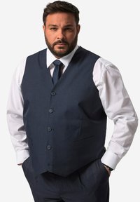 Marinblå väst med fem knappar och två fickor, bärs över en vit skjorta och kompletteras av ett mörkt slips. Slät textur.
