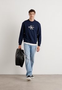 Sweatshirt azul-marinho com logótipo branco, jeans azul claro e bolsa preta. Estilo casual com um ajuste relaxado e design simples.
