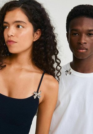 Two individuals wear clothing featuring decorative butterfly brooches. The woman wears a dark blue textured top; the man is in a white shirt.
