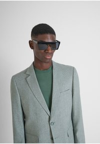 Blazer texturizado gris sobre un suéter verde, con solapa muesca y cierre de un solo botón. Unas gafas de sol negras grandes y angulares completan el look.