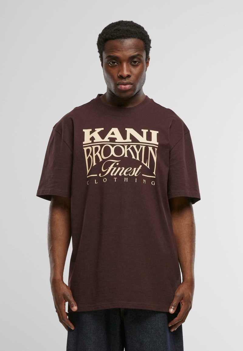 Dunkelbraunes Baumwoll-T-Shirt mit großem cremefarbenem Text: "KANI BROOKLYN Finest Clothing." Kurze Ärmel und lockere Passform.