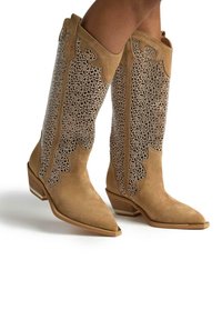 Botas de vaquero de ante marrón con punta afilada, que presentan intrincados adornos de cuentas, bordes ondulados y un tacón bajo y apilado.