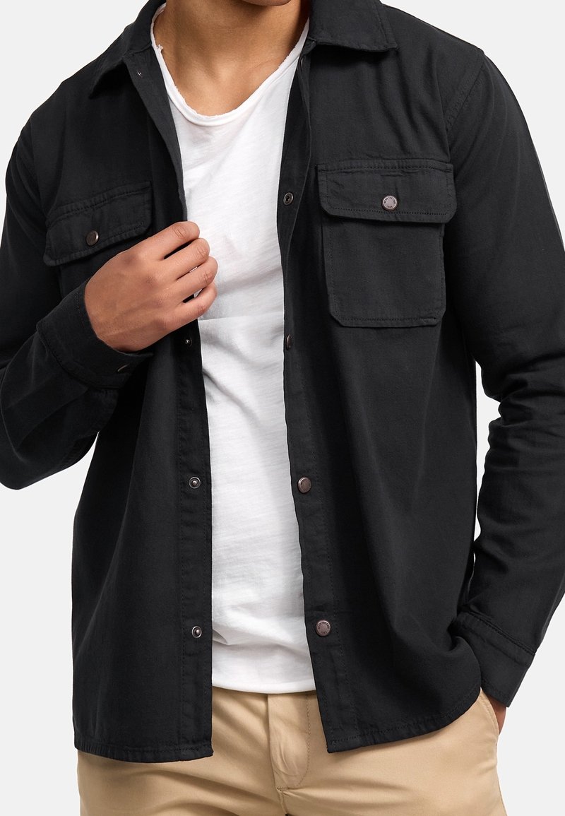 Homme portant une chemise noire boutonnée sur un t-shirt blanc et un pantalon beige, une main ajustant le col de la chemise et l'autre dans la poche.