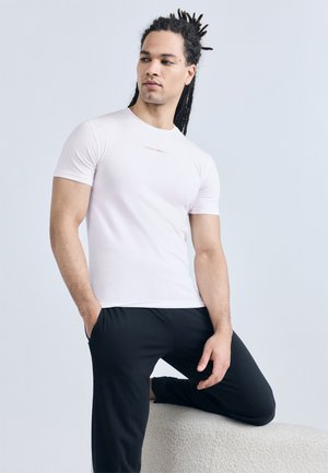 Uomo con dreadlocks che indossa una maglietta bianca aderente e pantaloni neri, inginocchiato su una gamba su una panchina testurizzata contro uno sfondo chiaro.