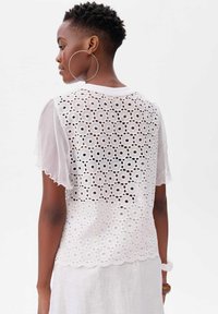 Biała bluzka z krótkim rękawem, ozdobiona skomplikowanym wzorem ażurowym, z falbaniastym dołem i lekkim materiałem. Posiada przezroczyste rękawy i okrągły dekolt.