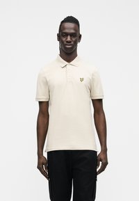 Polo color beige con colletto, maniche corte e un piccolo logo giallo a forma di farfalla sul petto, indossata con pantaloni scuri.