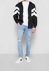 Černá lehká bunda s bílými chevronovými pruhy, doplněná o roztrhané světle modré džíny a bílé tenisky s černými doplňky.