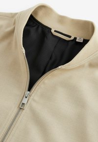 Veste beige avec fermeture éclair argentée partiellement ouverte, montrant une doublure intérieure en mesh noir et une petite étiquette de taille près du col.