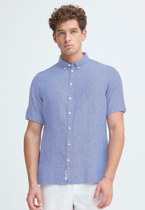 Homme aux cheveux bouclés portant une chemise à manches courtes bleu clair et un pantalon blanc, debout devant un fond uni clair.