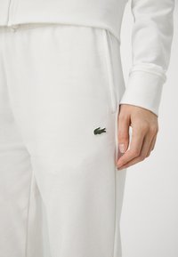 Jogging en coton blanc avec un petit logo de crocodile brodé vert sur le côté gauche. Le design comprend des poches latérales et des poignets élastiques.