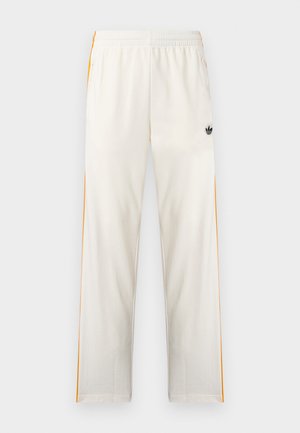 Λευκή φόρμα με πορτοκαλί ρίγες στο πλάι, ελαστική μέση, πλαϊνές τσέπες με φερμουάρ και μαύρο λογότυπο Adidas trefoil στο δεξί μηρό.