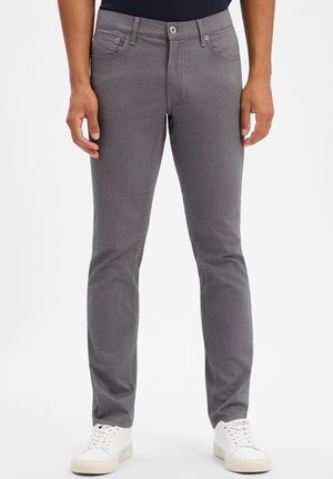 Mann trägt graue Slim-Fit-Hose und weiße Sneakers, steht vor einem einfarbigen weißen Hintergrund.