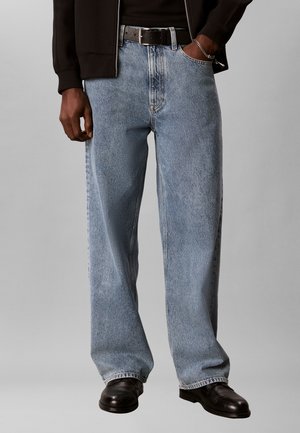 Mand iført lyseblå jeans med vide ben, sort bælte, sorte sko og en mørk jakke med den ene hånd i lommen mod en ensfarvet baggrund.
