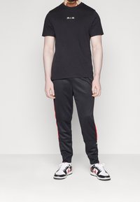 Musta t-paita, jossa on kultainen "AIR"-logo, yhdistettynä mustiin housuihin, joissa on punaiset sivuraidat. Kengissä on musta-valkoinen design.