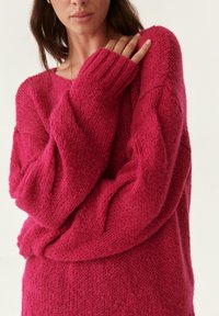 Suéter de punto rosa de gran tamaño con tejido texturizado, puños acanalados y un corte relajado. El detalle de las mangas muestra una caída suelta y acogedora.