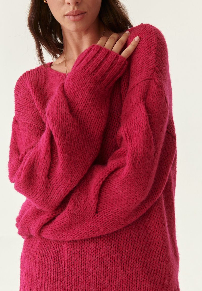 Suéter de punto rosa de gran tamaño con tejido texturizado, puños acanalados y un corte relajado. El detalle de las mangas muestra una caída suelta y acogedora.