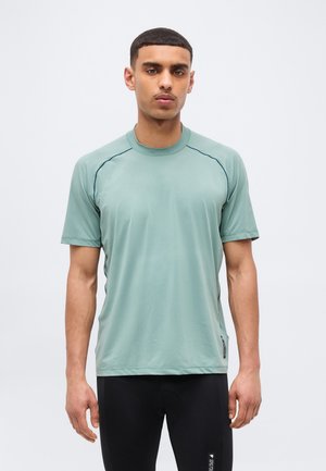 Νεαρός με κοντά μαλλιά που φοράει αθλητική κοντομάνικη πράσινη μπλούζα και μαύρο παντελόνι, στέκεται μπροστά σε απλό φωτεινό φόντο.