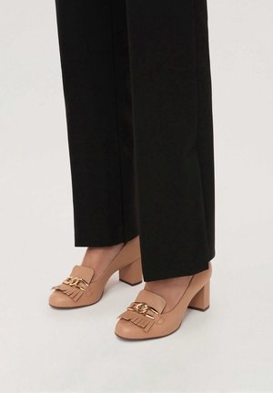 Chaussures beiges à talons blocs avec chaîne dorée et détails à franges portées avec un pantalon noir sur fond blanc.