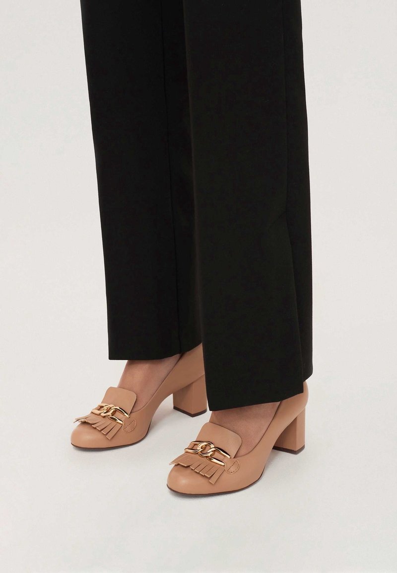Chaussures beiges à talons blocs avec chaîne dorée et détails à franges portées avec un pantalon noir sur fond blanc.