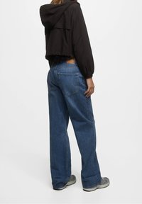 Kraftigt cropped sort jakke med hætte og pufærmer, parret med blå vidde jeans med baglommer og et lædermærke. Grå sneakers.