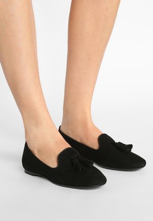 Voeten die zwarte suède loafers met tassels dragen, gezien vanuit iets bovenop tegen een effen lichte achtergrond.