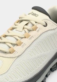 Világos színű sportcipő hálós felsőrésszel, texturált ráfedőkkel, bézs fűzőkkel és szürke talppal, jellegzetes mintázati részletekkel.