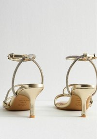 Ein Paar goldene, verstellbare High-Heelsandalen mit mit Strass besetzten Knöchelriemen und schmalen Absätzen, von hinten auf einer weißen Oberfläche gezeigt.