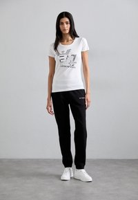 T-shirt bianca in cotone con stampa grafica, maniche corte e scollo rotondo. Abbinata a pantaloni jogger neri e sneakers bianche.