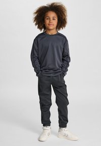 Barn med lockigt hår i en mörk idrottsoutfit och vita sneakers står självsäkert framför en neutral bakgrund.