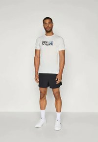 Vīrietis, kurš ģērbjas baltā New Balance t-kreklā, melnās šortos, baltās zeķēs un baltās sporta apavās, stāv uz vienkārša gaiši pelēka fona.