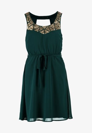 Femme portant une robe sans manches vert foncé avec un col orné de sequins dorés, des collants noirs transparents, des talons noirs, et tenant un sac à bandoulière noir avec chaîne.