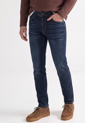 Homme portant un jean slim bleu foncé, des chaussures marron à lacets et une chemise marron à manches longues, debout avec une main dans la poche.